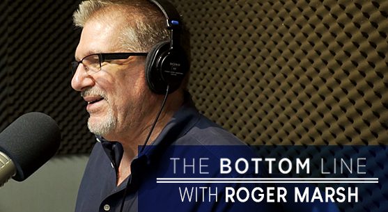 A man wearing glasses and headphones speaks into a microphone in a soundproof studio. Text overlay reads, "The Bottom Line with Roger Marsh.