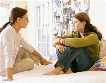 Two people sit on a bed facing each other, engaged in a serious conversation. One is wearing glasses and light clothing, while the other wears a green top and jeans. A collage of photos is visible on the wall behind them.
