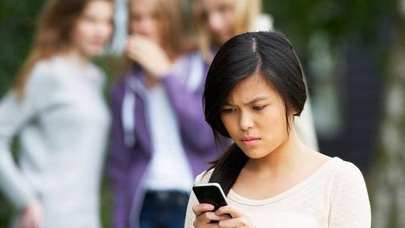 A teenage girl looks upset while reading something on her phone. In the blurred background, three other girls stand together, appearing to whisper and gossip.