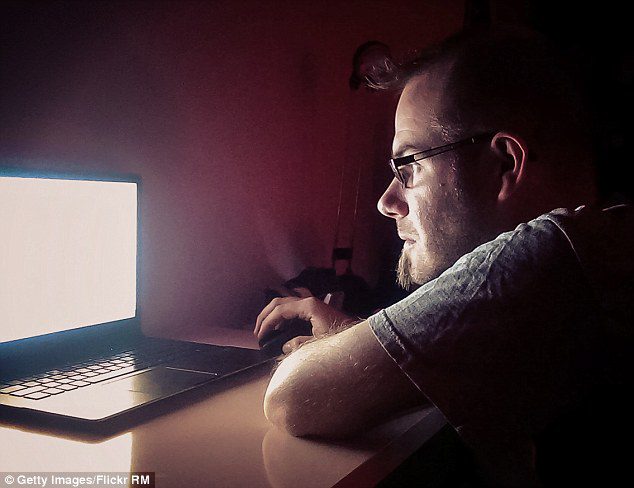 A man with glasses sits at a desk in a dimly lit room, illuminated by the bright screen of an open laptop as he uses a mouse.