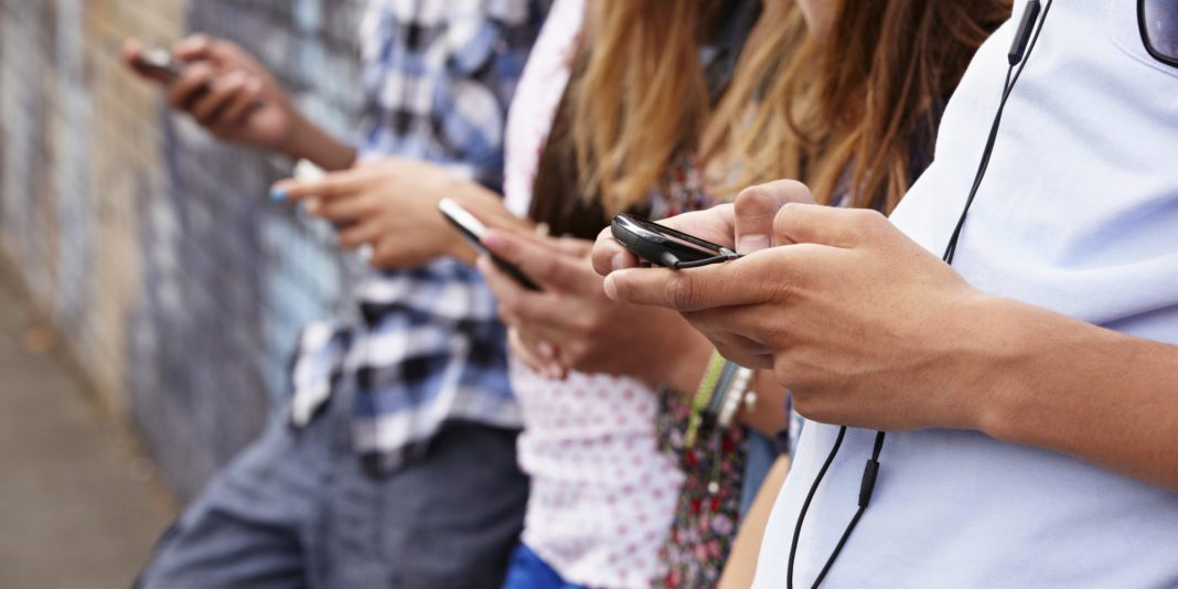 Three people stand side by side outdoors, each holding and looking at their smartphones. Their faces are not visible, and they are casually dressed. One person has earphones around their neck.