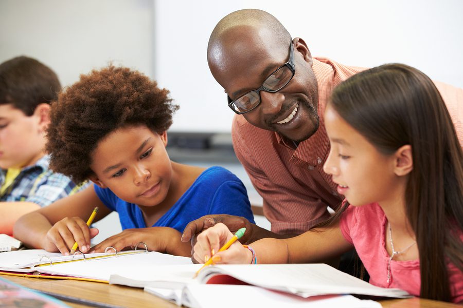 A teacher smiles and helps two students, a girl and a boy, with their schoolwork at a desk in a classroom. The students are writing in notebooks and appear to be engaged in learning.