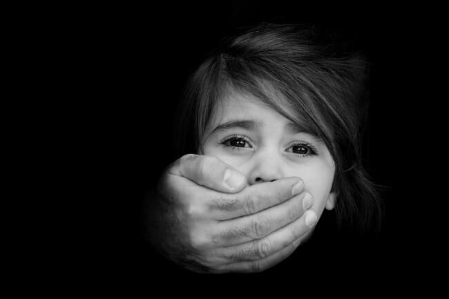 A young child with a fearful expression has a hand covering their mouth against a black background, suggesting distress or a need for help.
