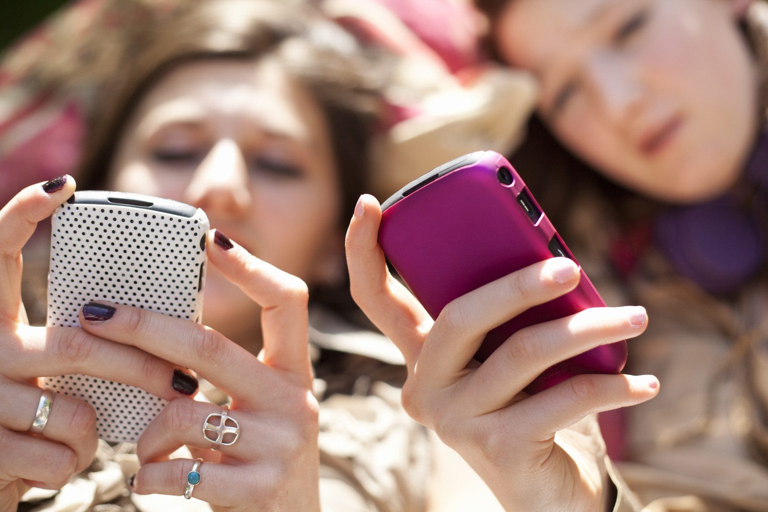 Two people lying down closely together, each focused on their smartphone. One phone has a white textured case, and the other has a bright pink case. Both individuals are blurred, with their hands and phones in sharp focus.