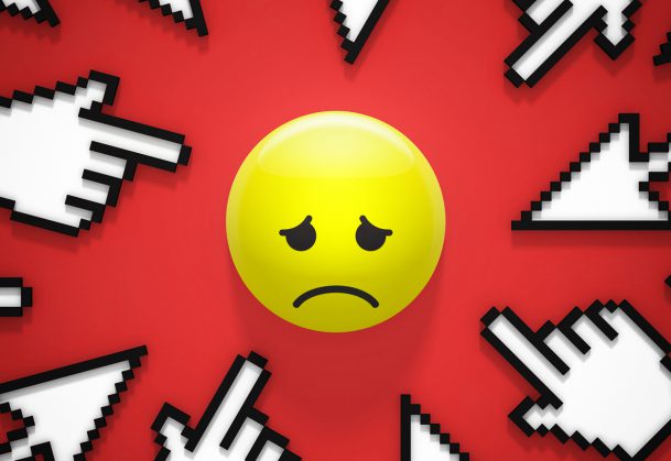A sad yellow emoji is surrounded by several pixelated computer cursor icons pointing toward it on a red background, symbolizing online bullying or harassment.