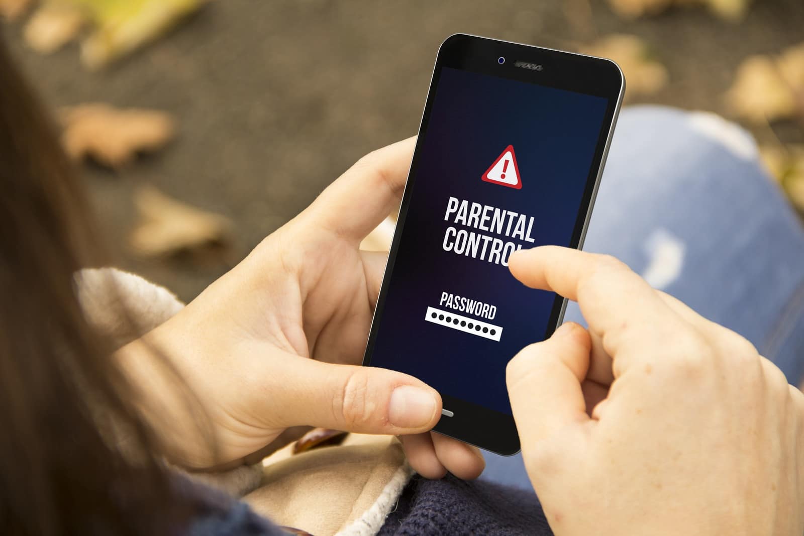 A person holds a smartphone showing a parental control screen with a password entry field and warning symbol, seated outdoors with fallen leaves in the background.