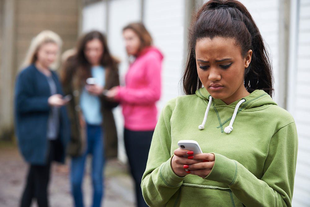 A teenage girl in a green hoodie looks upset while using her phone. In the blurred background, three other girls stand together, also looking at their phones.