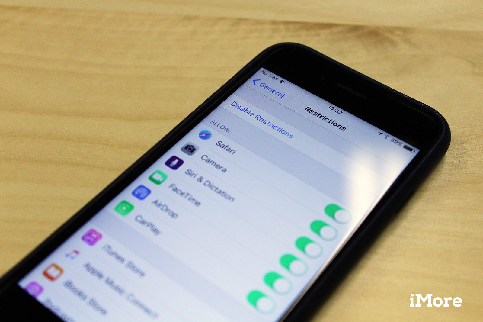 A smartphone displaying the 'Restrictions' settings screen, showing options to enable or disable access to apps like Safari, Camera, Siri & Dictation, and FaceTime; the phone rests on a wooden surface.