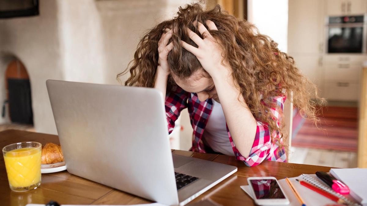 A person with curly hair sits at a table, holding their head in frustration while looking at a laptop. A glass of orange juice, a croissant, phone, and notebooks are on the table.