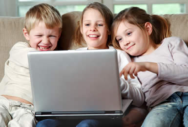 Three smiling children sit close together on a couch, looking at a laptop screen and appearing to enjoy what they see. One child is pointing at the screen, and all seem engaged and happy.