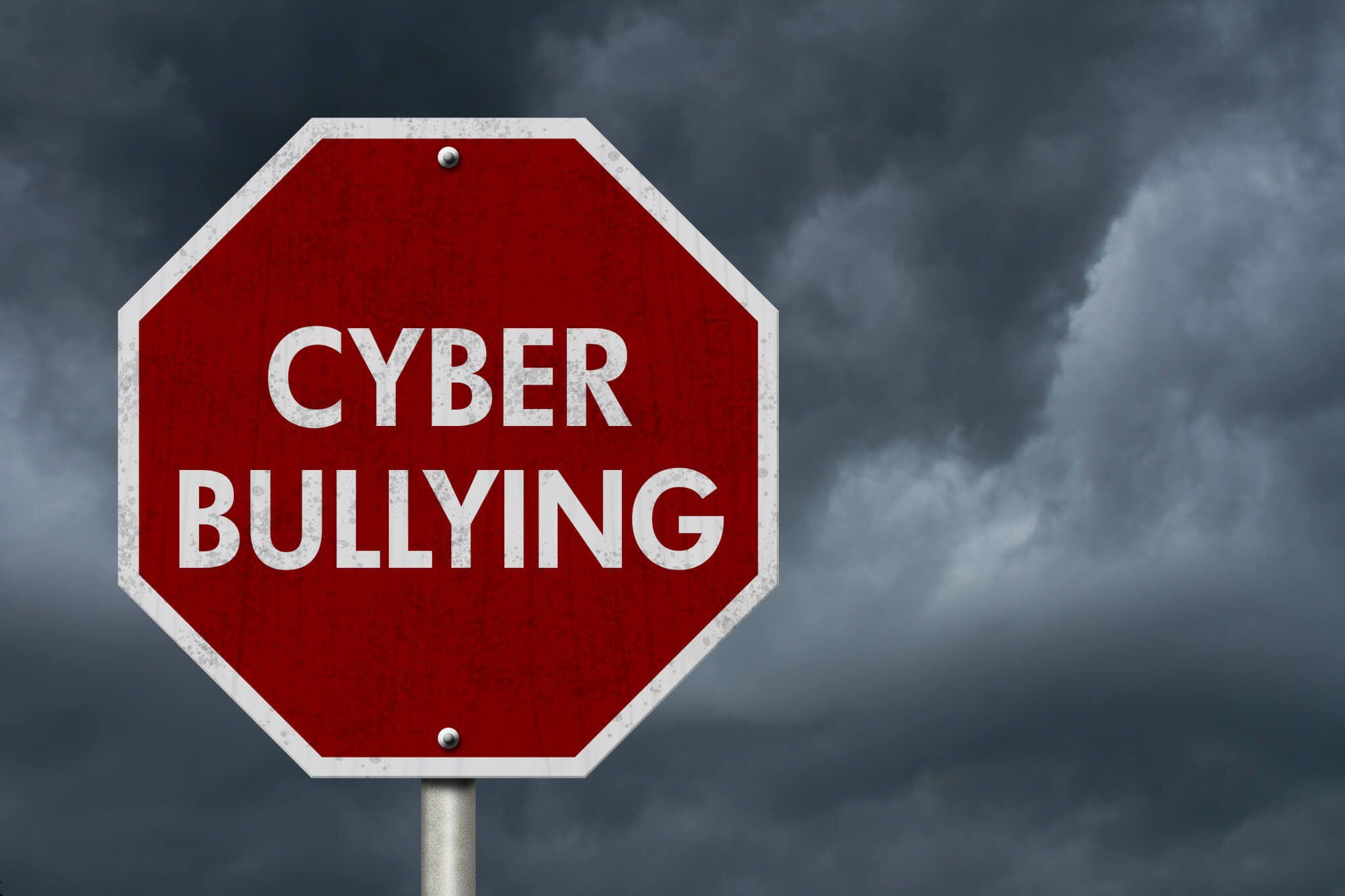 A red stop sign with the words “CYBER BULLYING” in white capital letters stands against a dark, cloudy sky, symbolizing a warning or call to stop cyberbullying.