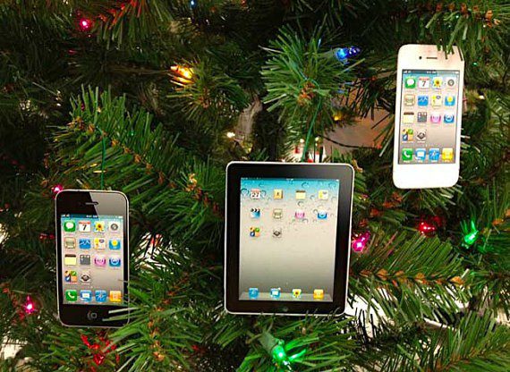 Three Christmas tree ornaments shaped like smartphones and a tablet hang on a green Christmas tree decorated with colorful lights. The devices display home screens with app icons.