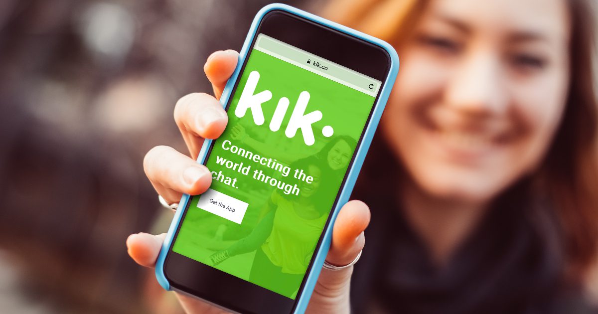 A person holds a smartphone close to the camera showing the Kik app website with the text "Connecting the world through chat" on a bright green screen. The person is smiling in the blurred background.