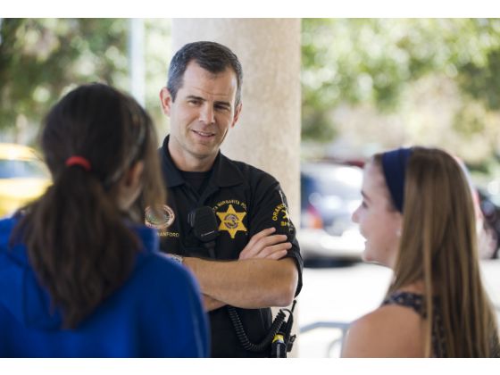 A police officer in uniform stands outside with arms crossed, smiling and talking to two teenage girls. The background is blurred with trees and parked cars visible.