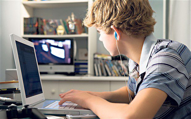 A teenage boy with blond hair sits at a desk using a laptop, wearing blue earphones. He is looking at the computer screen, with shelves of books and DVDs visible in the background.
