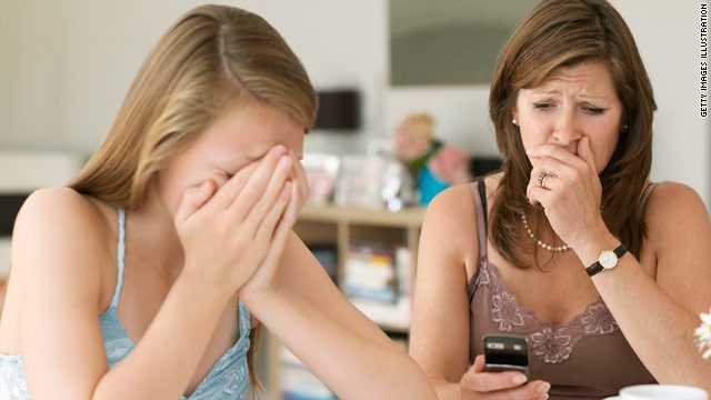 A teenage girl covers her face with her hands while an older woman beside her looks worriedly at a phone, holding her hand to her mouth. They are sitting together at a table in a home setting.