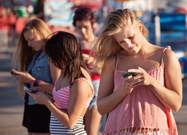 Four young women stand outdoors in casual summer clothes, each focused on their smartphones, seemingly unaware of their surroundings. The background is blurred, suggesting an outdoor event or fair.
