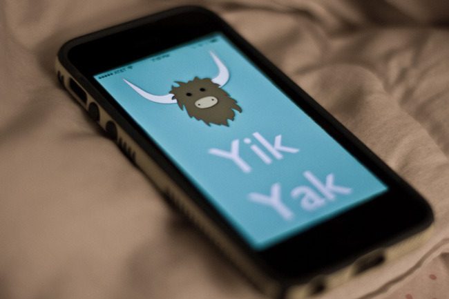 A smartphone on a bed displays the Yik Yak app logo, featuring a cartoon yak head and the words "Yik Yak" on a light blue background.