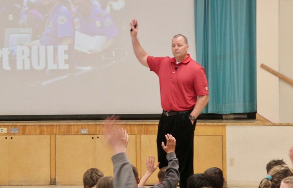A man in a red polo shirt stands in front of a projected screen, holding up one hand. Several students in the audience raise their hands. The setting appears to be a school or auditorium.