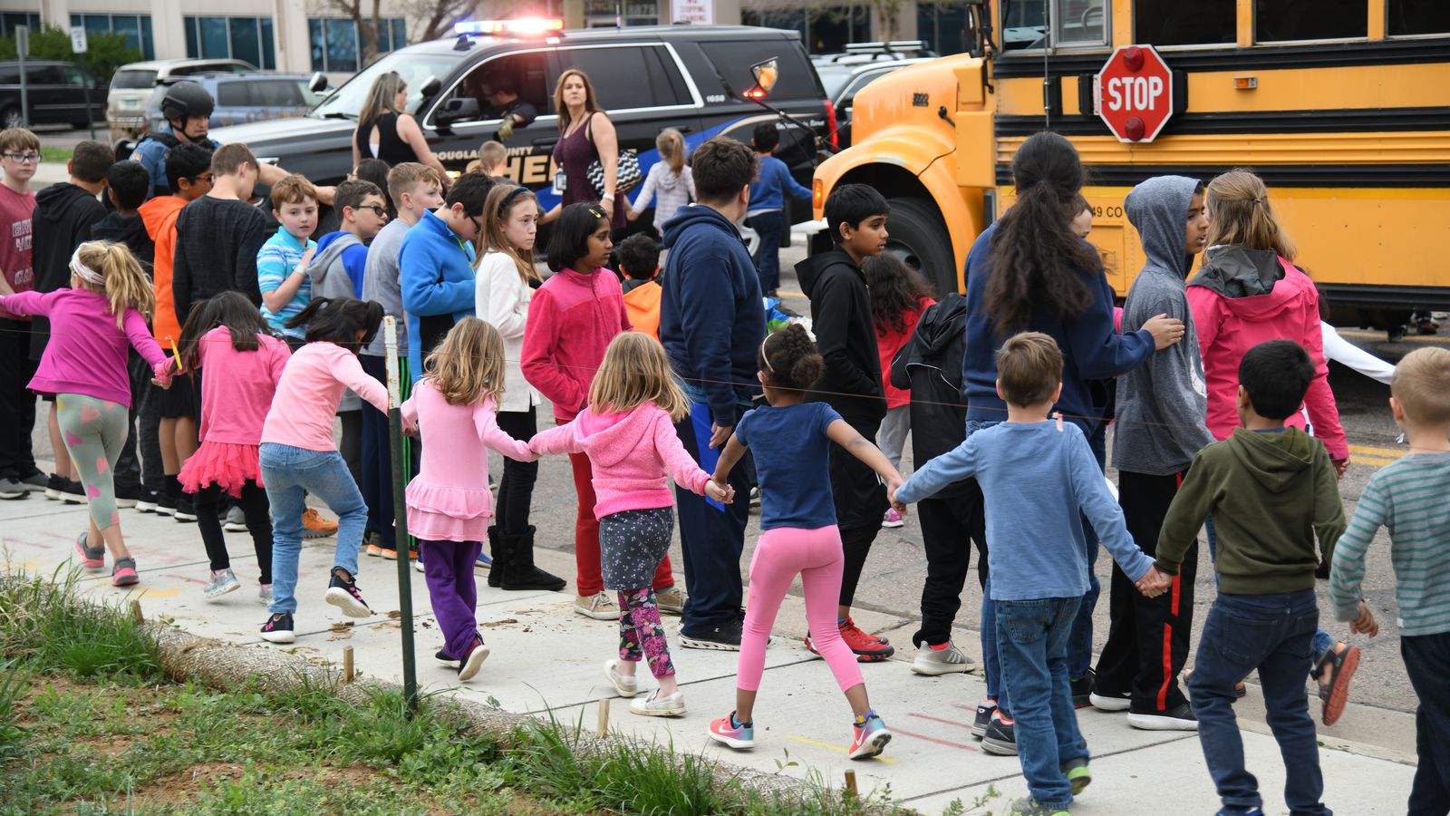 A large group of children, some holding hands, are being led away from a yellow school bus near a sidewalk, with adults and a sheriff’s car nearby. The scene appears orderly and supervised.