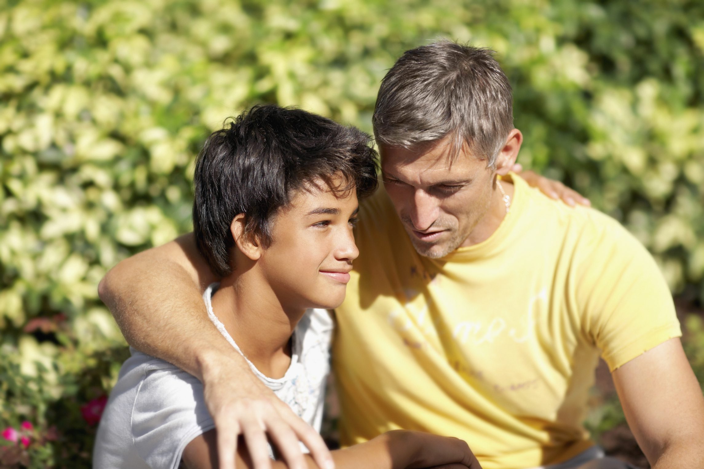 A man and a teenage boy sit close together outdoors, both smiling gently. The man has his arm around the boy’s shoulders. Green foliage is blurred in the background, suggesting a garden or park setting.