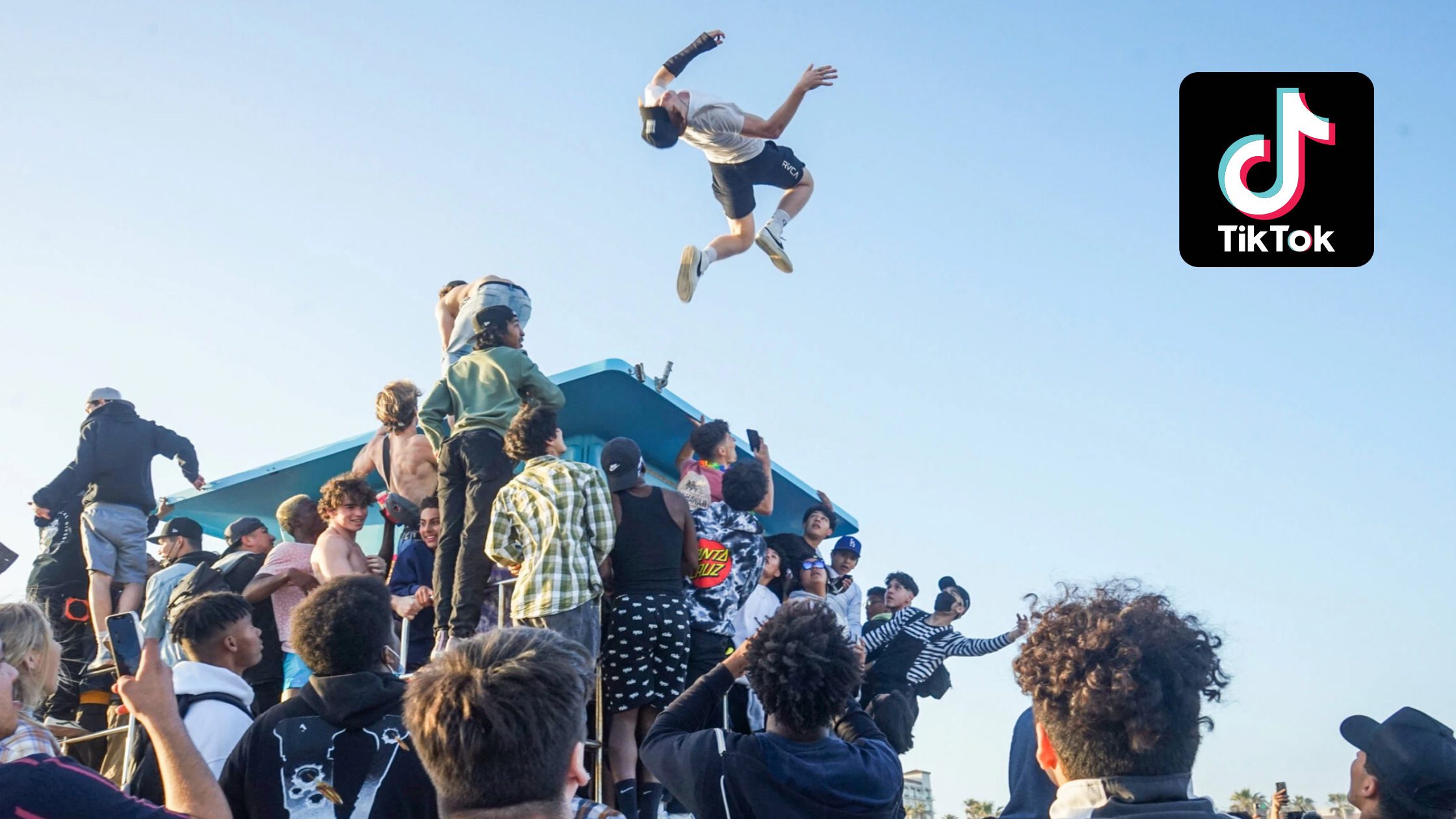 A crowd gathers around a lifeguard tower as one person is thrown high into the air. The sky is clear and the TikTok logo is visible in the top right corner.