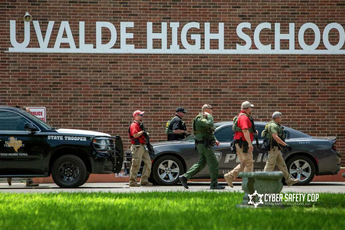 Law enforcement officers walk in front of Uvalde High School, with police vehicles parked nearby. The school name is visible on the building behind them.