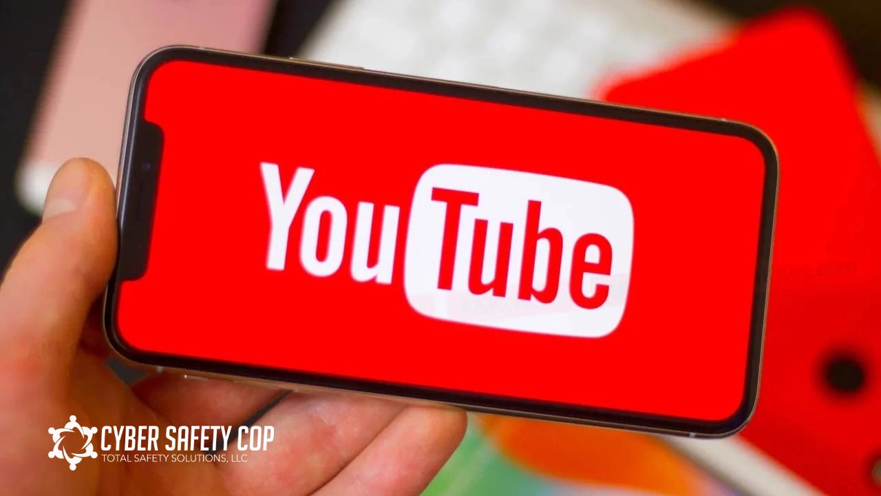 A hand holds a smartphone displaying the YouTube logo on the screen. The background is blurred with colorful objects, and a "CYBER SAFETY COP" logo appears in the lower left corner.