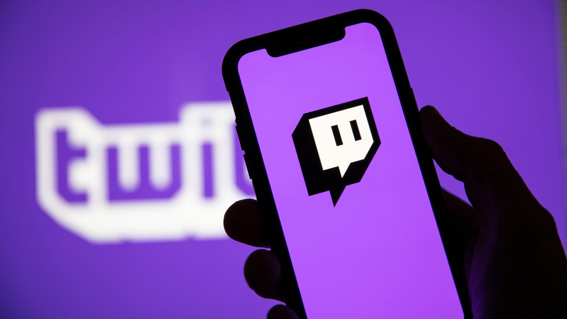 A hand holds a smartphone displaying the Twitch logo on its screen, with a larger blurred Twitch logo in the purple background.