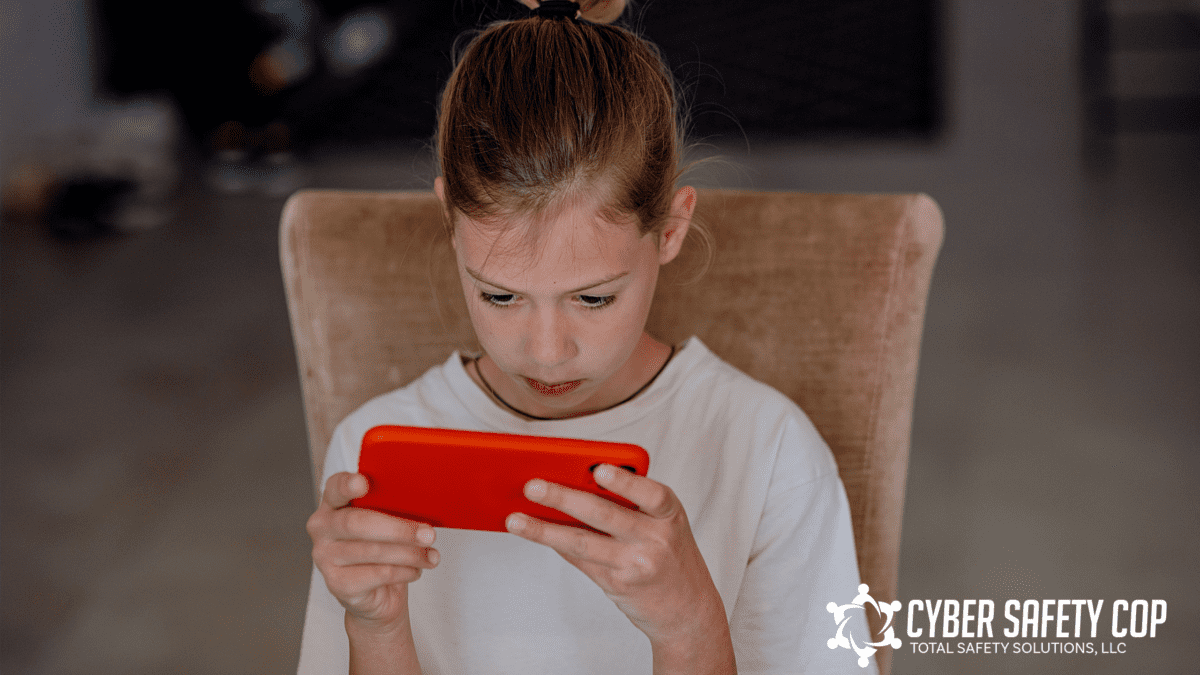 A young girl sits on a tan chair, intently looking at a red smartphone. The Cyber Safety Cop logo appears in the bottom right corner.