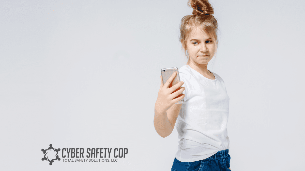 A young girl with blonde hair in a bun holds a smartphone and looks at the screen. She wears a white t-shirt and blue jeans. The logo “Cyber Safety Cop, Total Safety Solutions, LLC” is in the corner.