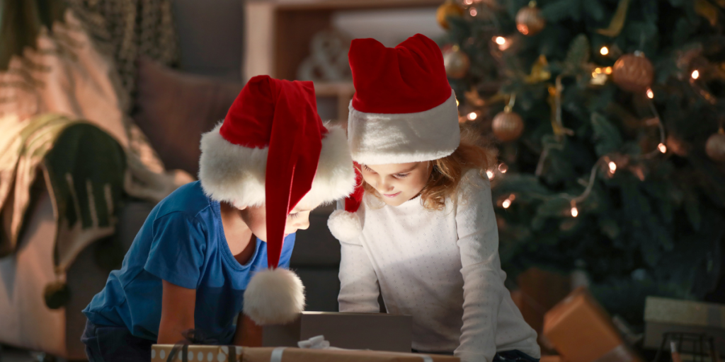 Two children wearing Santa hats sit together by a Christmas tree, looking at a glowing tablet screen. Surrounded by gifts and festive decorations, they enjoy their Christmas while learning about staying Kids Safe Online with their new gadgets.