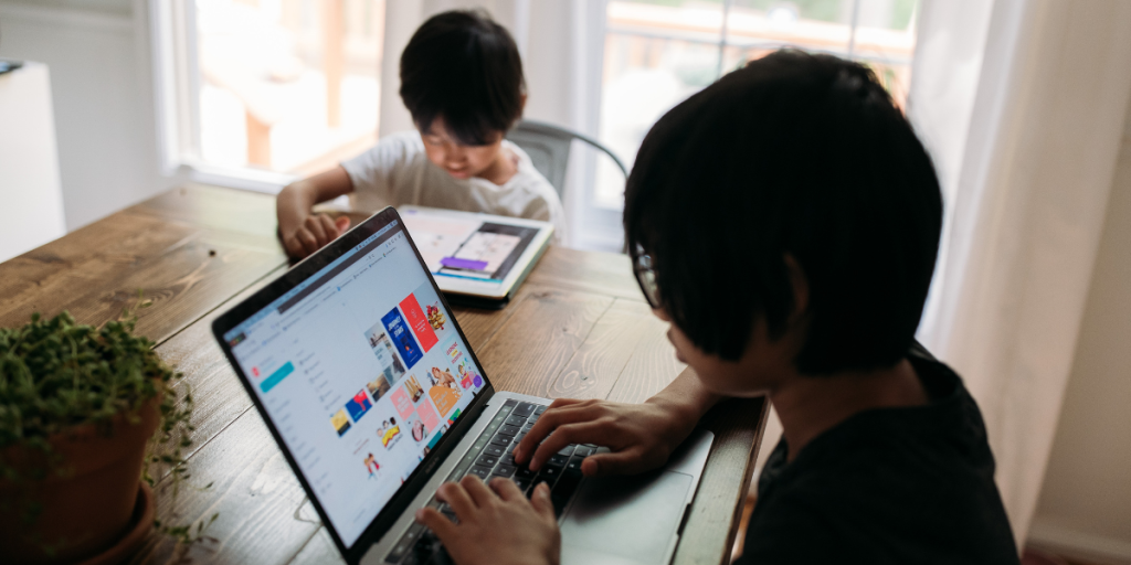 Two kids sit at a wooden table near a window. One uses a laptop while the other engages with a tablet. A potted plant is on the table, and natural light fills the room as parents watch nearby.