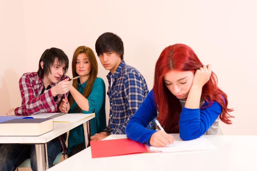 Four students sit in a classroom. Three are whispering, possibly discussing cyberbullying, while the fourth—who has red hair and sits alone—appears sad or left out, focused on writing.