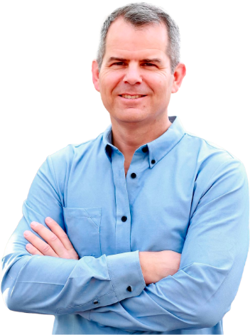 A man with short gray hair, wearing a light blue button-up shirt, stands with his arms crossed and smiles at the camera, holding a book against a plain white background.