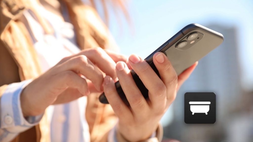 A person uses a smartphone outdoors with blurred buildings in the background. In the foreground, an Apple Hot Tub app icon featuring a bathtub is visible, highlighting modern wellness and relaxation technology.