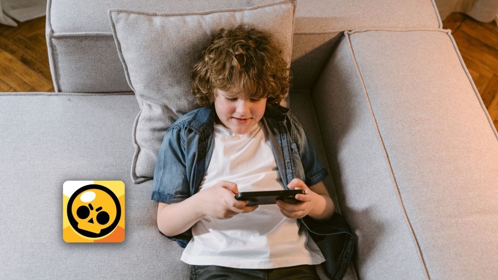 A young boy with curly hair lies on a light gray couch, smiling while playing on a smartphone. The Brawl Stars game app icon and exciting features are visible in the bottom left corner of the image.