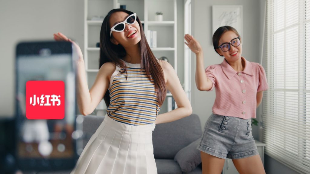 Two young women dance and smile in a living room, recording themselves on a smartphone. Highlighting RedNote’s features, the Xiaohongshu logo appears on the left side of the image as they stay mindful of cyber internet safety.
