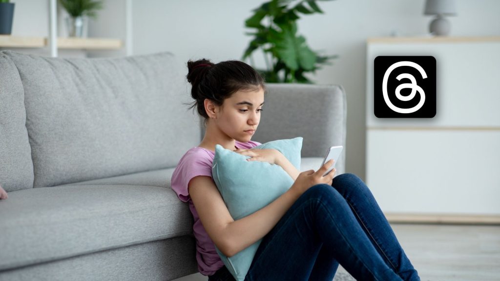 A young girl sits on the floor by a couch, holding a pillow and looking at her smartphone. The Threads app logo, known for its features focused on cyber internet safety, is visible on the wall behind her.