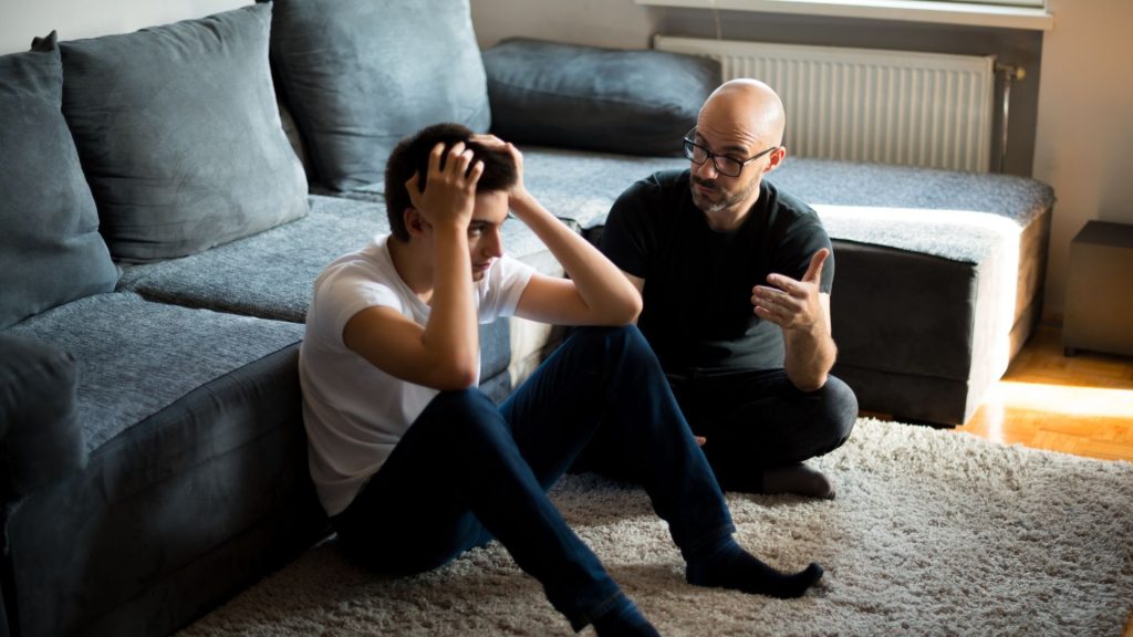 A young man sits on the floor with his head in his hands, looking stressed, while an older man beside him offers advice—demonstrating parenting that fosters emotionally intelligent kids—in a living room with a gray sofa.