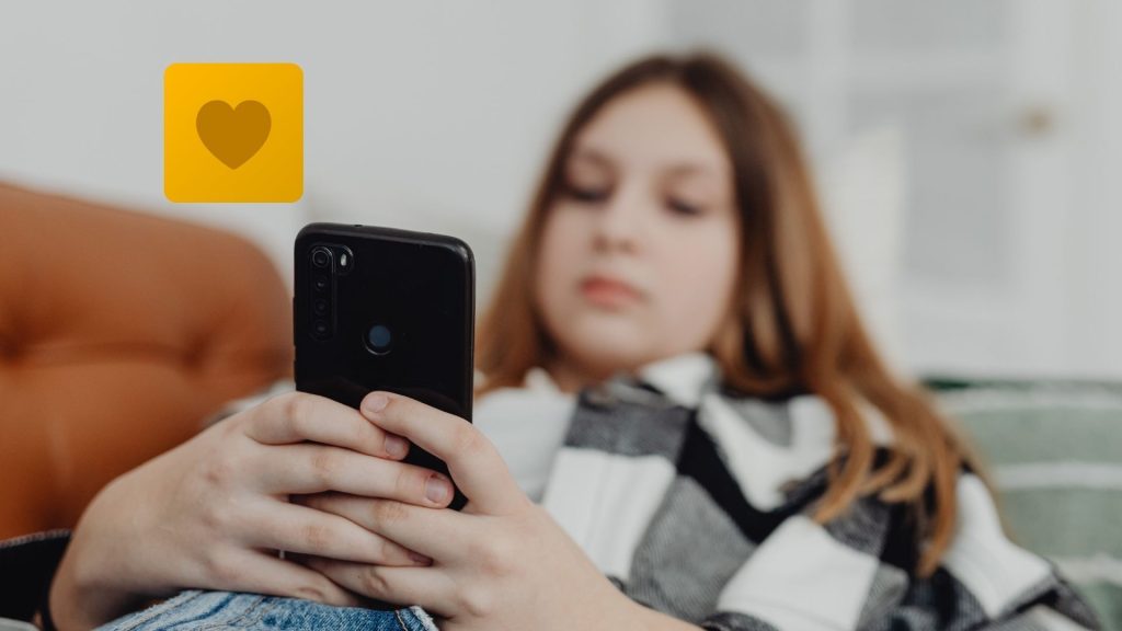 A young person reclines on a sofa, holding a smartphone. A yellow icon with a heart symbol appears near the phone, suggesting Locket App activity and sparking parents' interest in this popular social media platform.